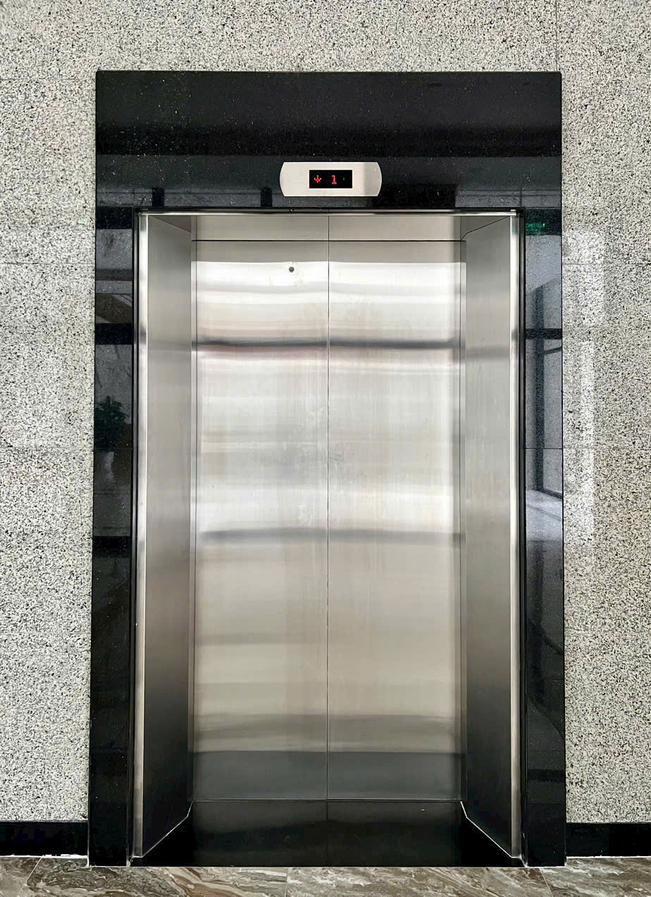 Cửa thang máy inox đóng kín với bảng hiển thị điện tử mũi tên xuống và số tầng